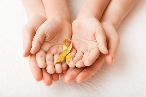 An adult cradles a child's hands in theirs, both holding a gold ribbon symbolizing childhood cancer