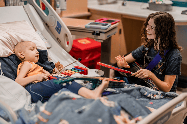 A young girl in a hospital bed plays musical instruments with a music therapist