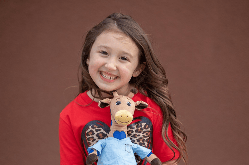 A young girl smiles as she holds a George the Giraffe stuffed toy
