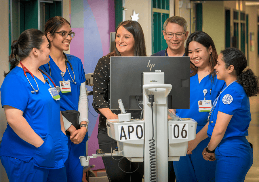 A nurse stands at a mobile computer station in a unit and speaks with nursing students