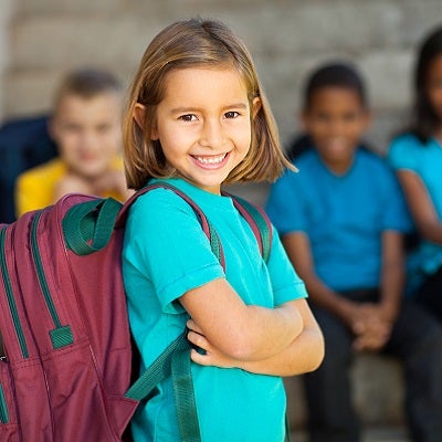 A young girl smiles as she stands by her school with her backpack