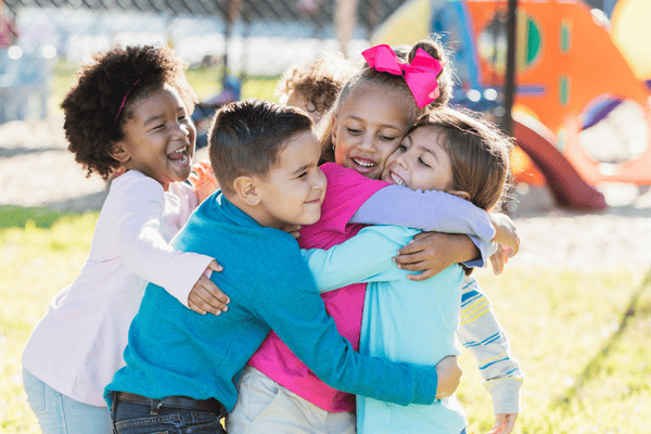 A diverse group of young children hug each other in front of a playground structure