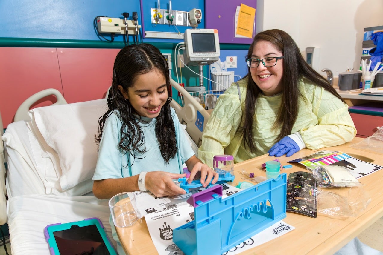 a girl plays at her hospital bed table with toys along with a Child Life Specialist