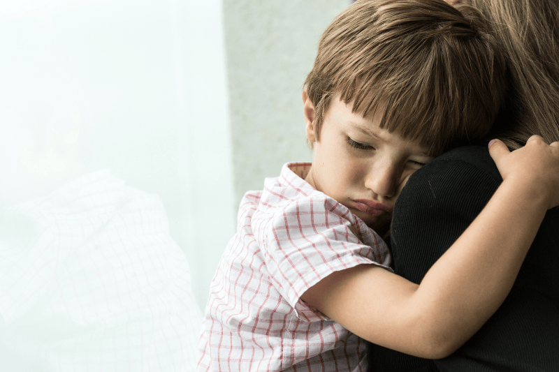 A tired young boy frowns as he is held by his mother