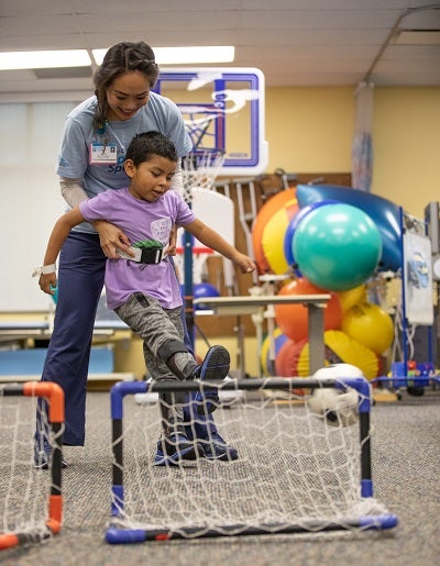 Photo of physical therapist helping young boy kick a soccer ball into net