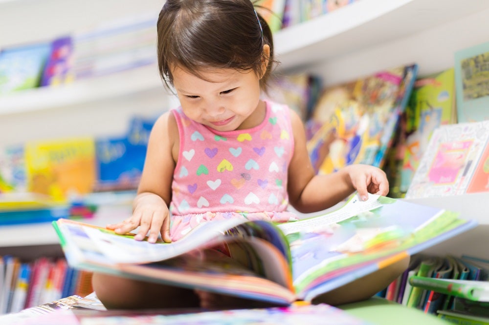 A young girl sits in a library and smiles as she reads a picture book